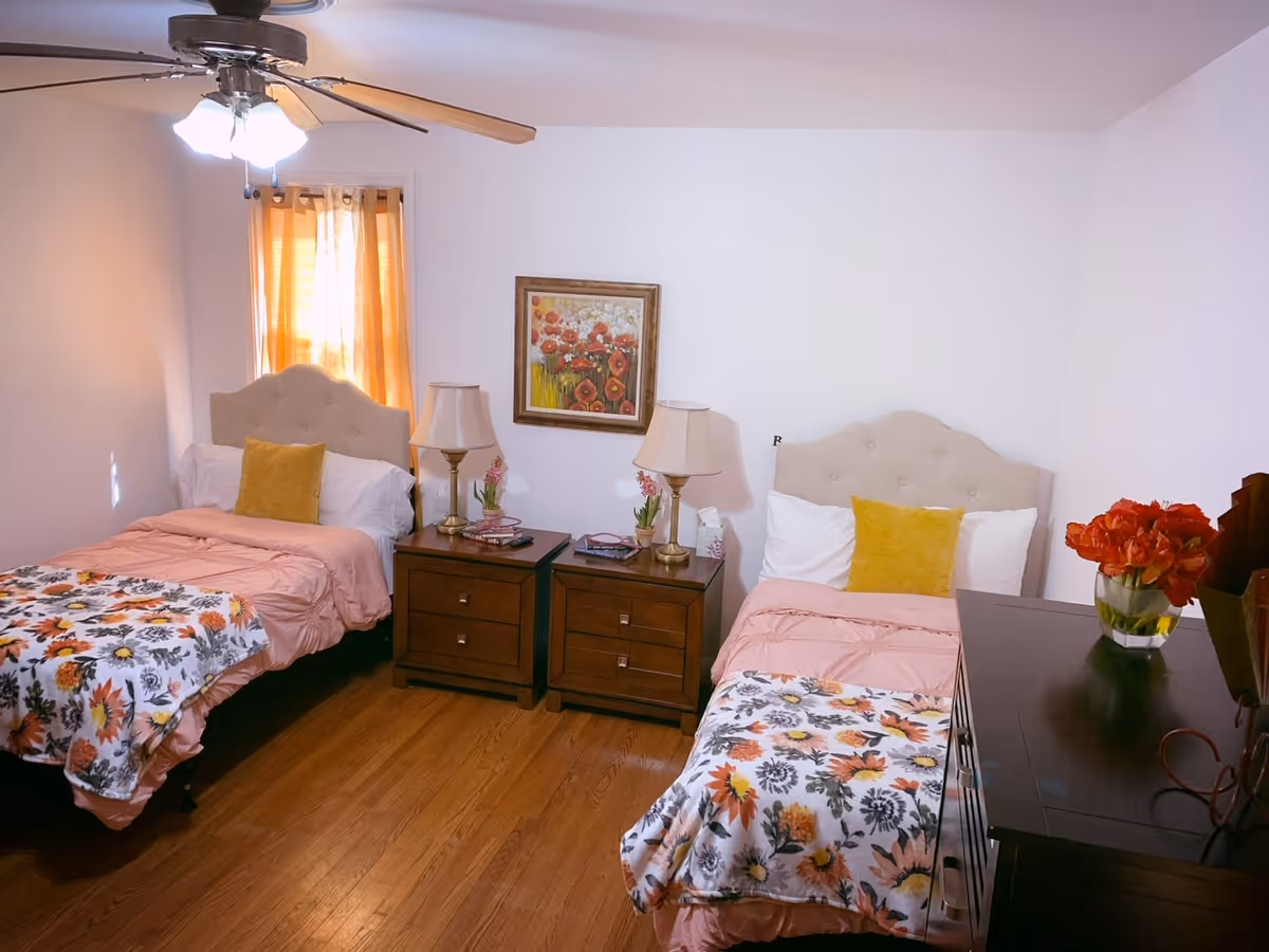 Bedroom with two twin beds, matching headboards, bedside tables with lamps, floral blankets, a dresser with flowers, and a ceiling fan.