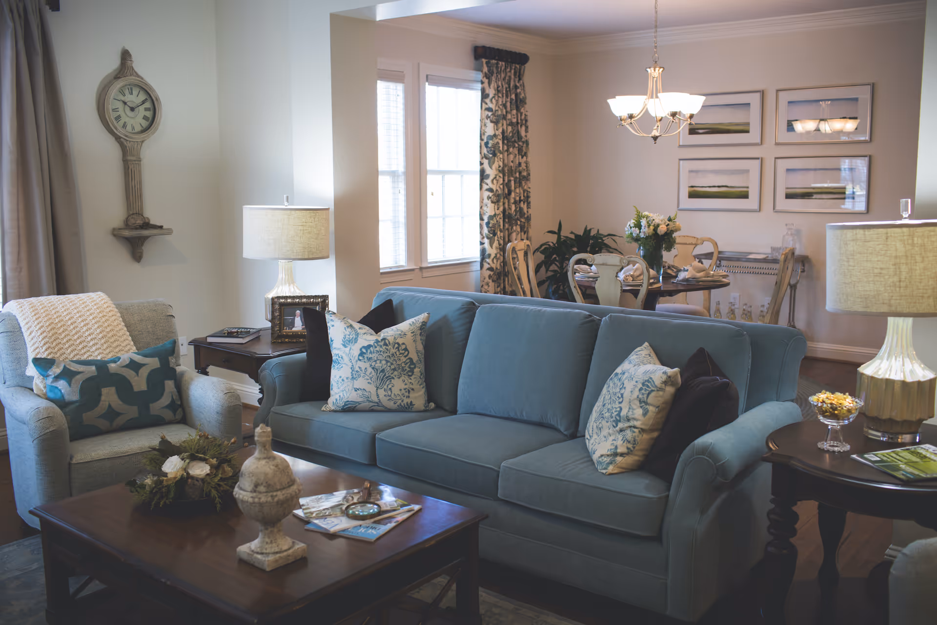 A cozy living room with a blue sofa adorned with patterned pillows, a matching armchair with a decorative pillow and throw blanket, a wooden coffee table with decorative items, and two side tables each with a lamp. In the background, there is a dining area with a round table set for four, floral curtains, and framed landscape pictures on the wall.