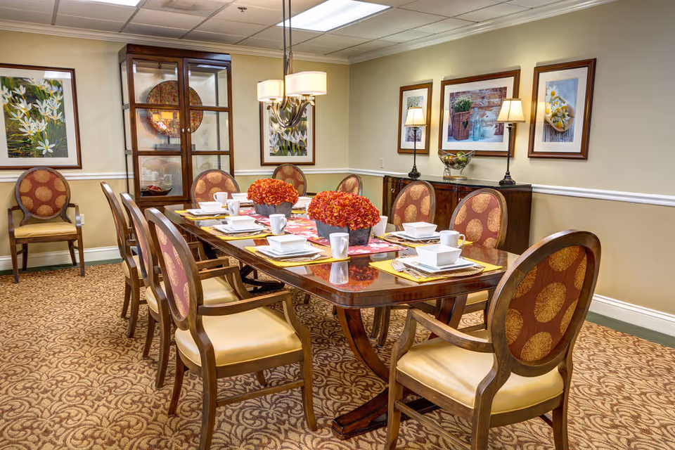 A formal dining room with a long wooden table set for eight people. The table is decorated with two centerpieces of red flowers and place settings including white cups, bowls, and plates on yellow placemats. The room has patterned carpet, beige walls with white trim, framed floral artwork, a wooden cabinet with glass doors, and two table lamps on a sideboard.