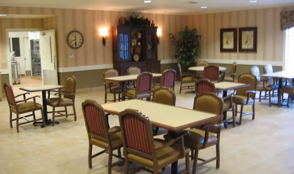 A dining room with several tables and chairs arranged neatly. The room has beige and green walls with striped wallpaper, a clock on the wall, a dark wooden cabinet with decorative plates, two framed pictures, and a potted plant in the corner. The floor is tiled, and there is a doorway leading to another room.