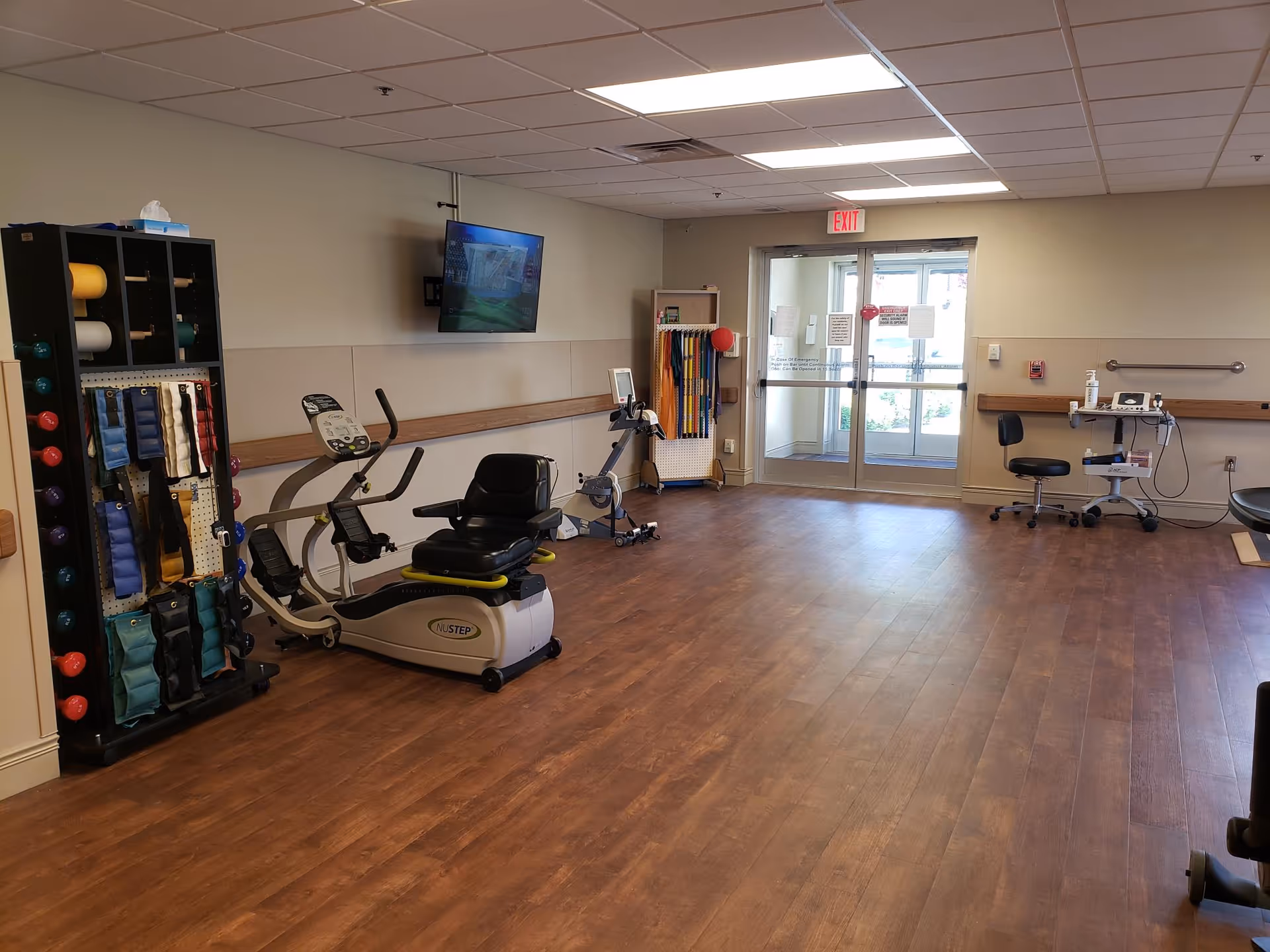 A spacious interior therapy/exercise room with recumbent exercise bikes, racks of weights and resistance bands, wood-look flooring and double glass exit doors.