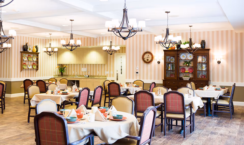 A well-lit dining room with multiple round tables covered with beige tablecloths, each set with colorful bowls, plates, cups, and folded napkins. The room features striped wallpaper, wooden flooring, and several chandeliers hanging from the ceiling. There is a wooden cabinet with decorative plates and plants, and a wall clock on the far wall.