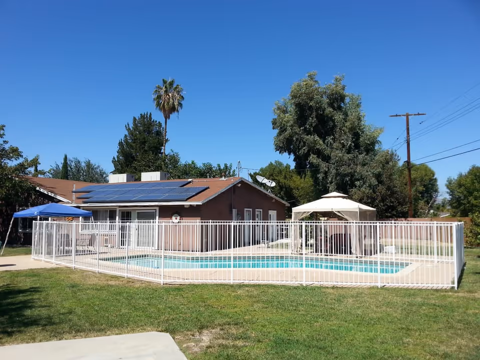 Outdoor view of a residential facility with a fenced swimming pool, a brown building with solar panels on the roof, a white canopy, and green trees under a clear blue sky.