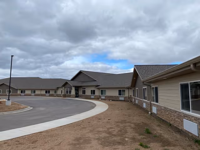 Exterior view of a single-story assisted living facility building with beige siding and stone accents, a curved driveway, and a cloudy sky overhead.
