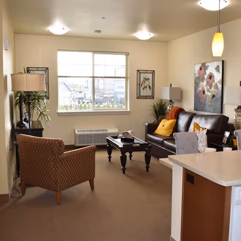A cozy living room in a senior living facility with a brown patterned armchair, a dark brown leather sofa with yellow and orange cushions, a wooden coffee table, two table lamps, a large window letting in natural light, and decorative artwork on the walls.