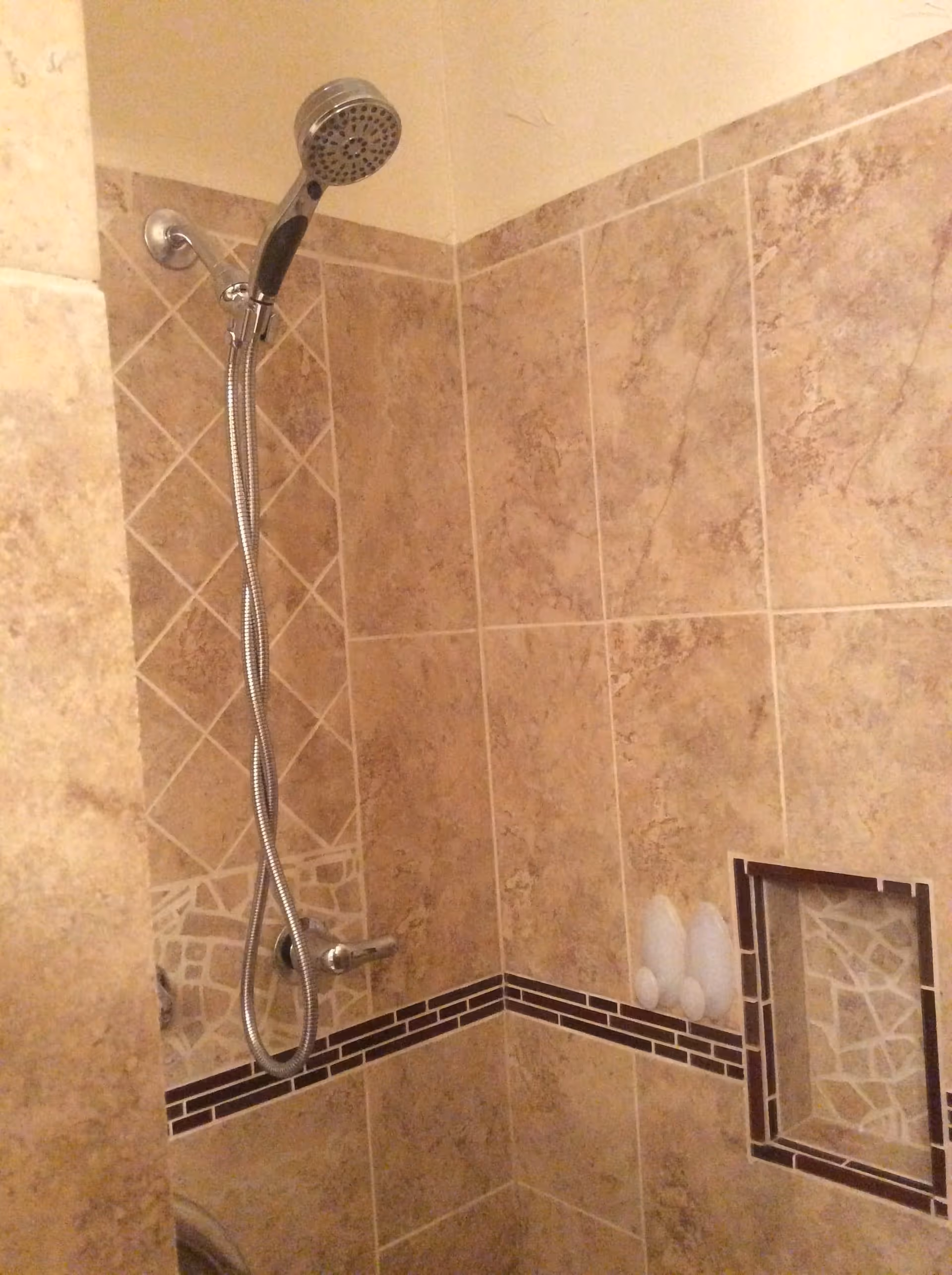 A tiled shower area with a handheld showerhead attached to a flexible hose mounted on the wall. The tiles are beige with a decorative horizontal strip and a built-in niche containing three bottles.