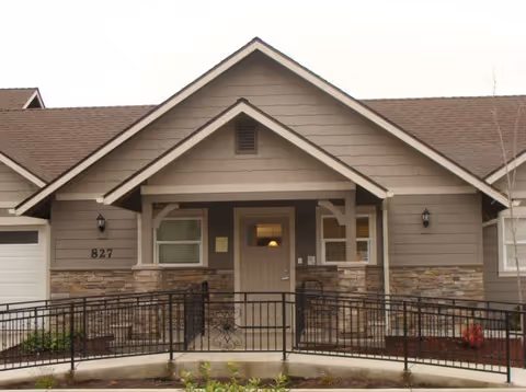 Front exterior view of a single-story residential building with a gabled roof, stone and wood siding, a central door with a small window, two windows on either side, and a black metal wheelchair ramp leading to the entrance. The building number 827 is visible on the left side near the garage door.