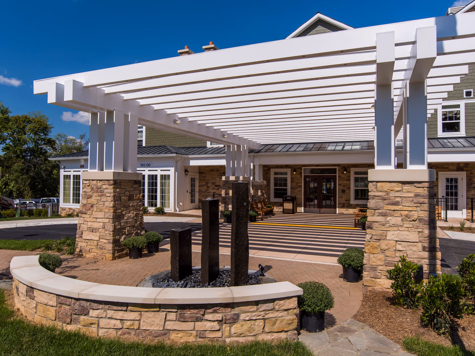 Entrance area of Brightview Great Falls Assisted Living & Memory Care featuring a stone and white pergola structure over a circular water fountain with three vertical black stone pillars. The building facade has stone and beige siding with large windows and a double glass door entrance under a covered walkway. Green shrubs and potted plants surround the area under a clear blue sky.