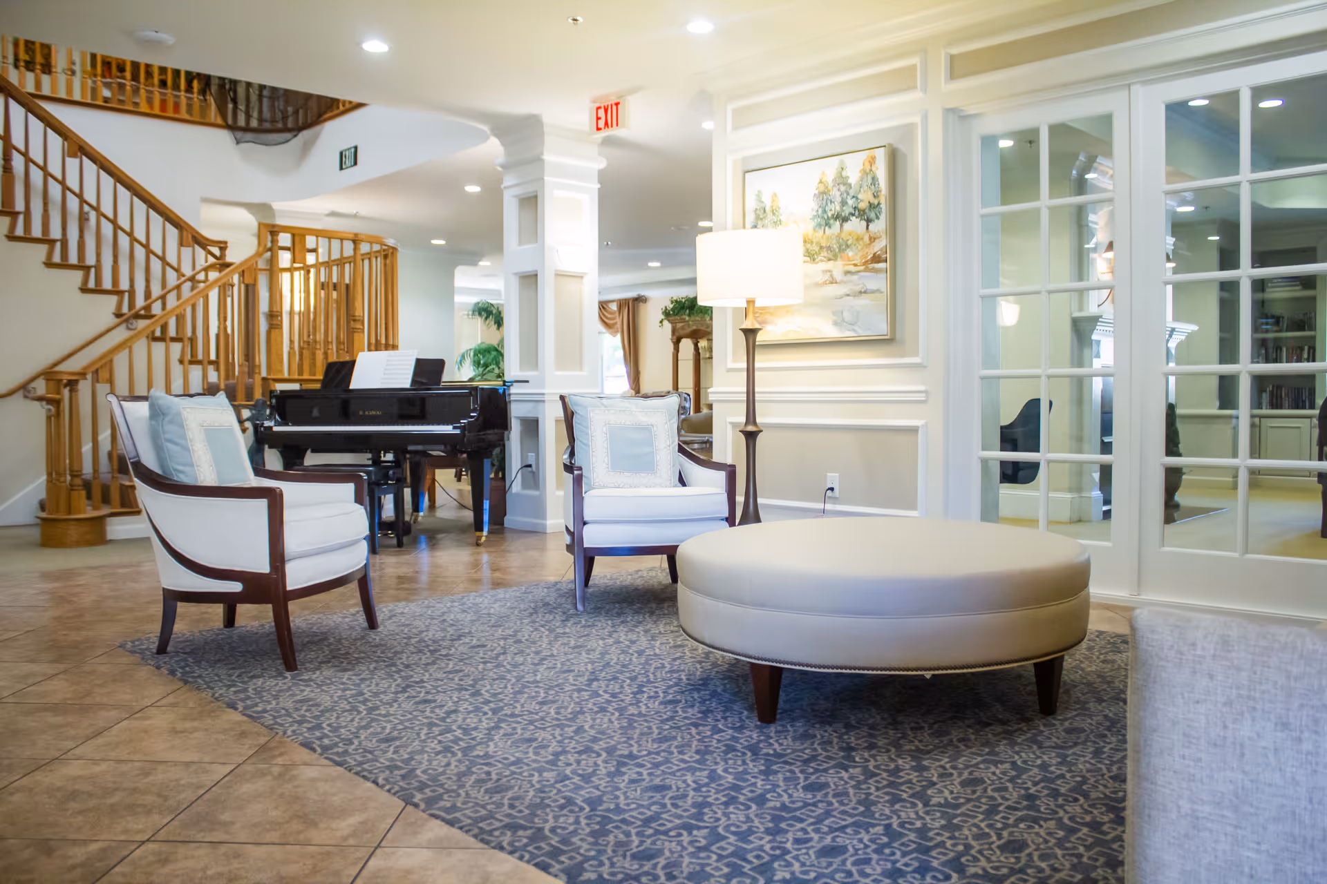 Bright communal lounge with upholstered chairs, a round ottoman, a piano, and a wooden staircase.