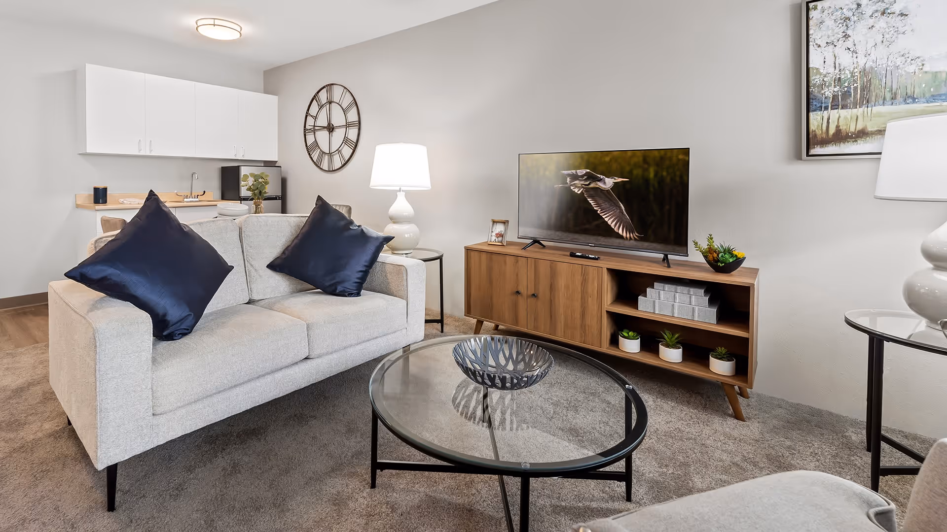 A modern living room with a light gray sofa adorned with two dark blue pillows, a round glass coffee table with a decorative bowl, a wooden TV stand with a flat-screen TV displaying a bird in flight, and a small kitchenette in the background with white cabinets and a large wall clock.
