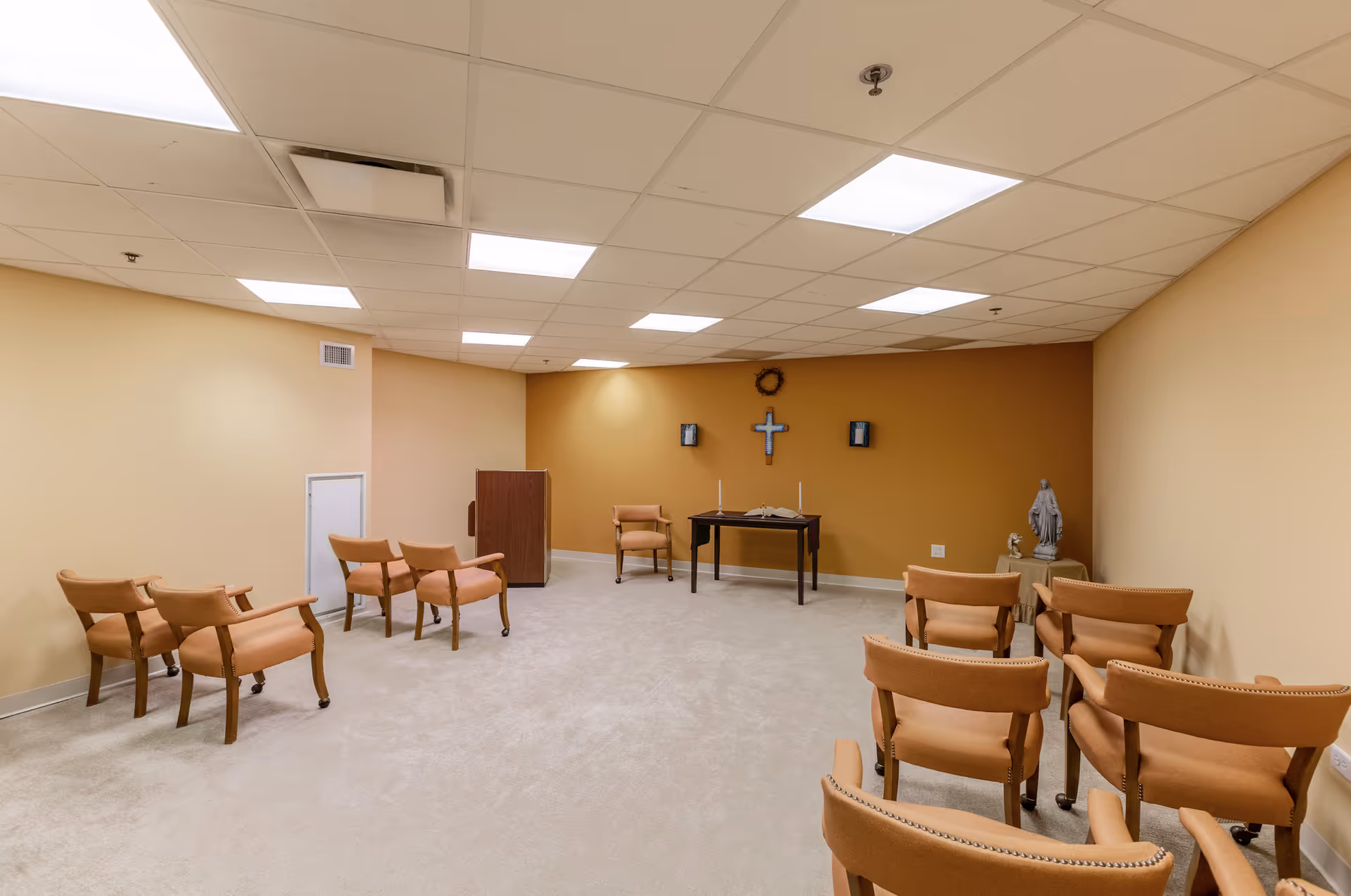 A small chapel room with beige walls and carpeted floor, featuring several rows of wooden chairs with padded seats facing a small table with an open book, two candles, and a cross mounted on the wall behind it. There is also a podium and a statue on a side table in the corner.