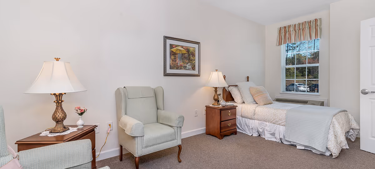 A cozy bedroom in a senior living facility featuring a single bed with white and patterned bedding, a wooden nightstand with a lamp and alarm clock, a comfortable upholstered armchair, a framed painting on the wall, and a window with striped valance showing a view of parked cars outside.
