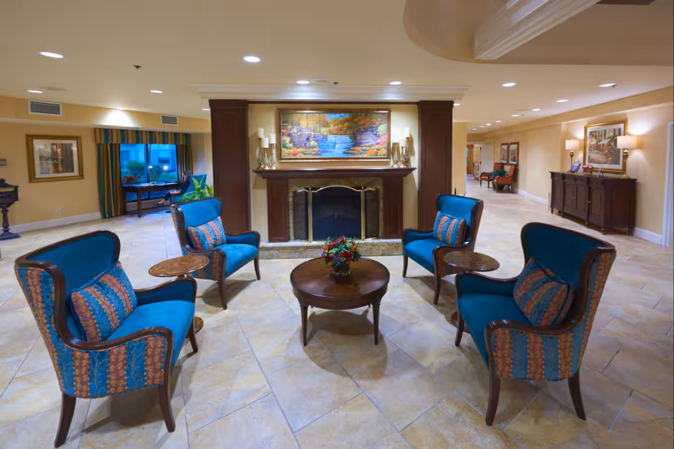 A cozy seating area in a senior living facility featuring four blue upholstered armchairs with patterned cushions arranged around a round wooden coffee table with a floral centerpiece. Behind the seating area is a fireplace with a colorful landscape painting above it. The room has tiled floors, warm beige walls, recessed lighting, and additional furniture and artwork along the walls.