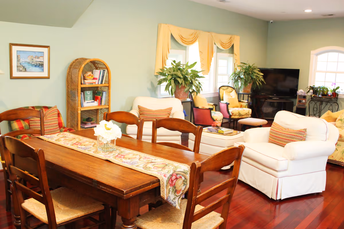 Cozy senior living lounge featuring a wooden dining table, white sofas, colorful chairs, and potted plants.