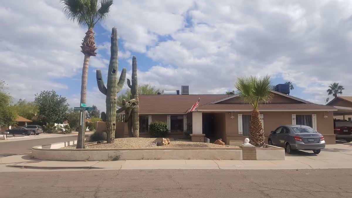 Single-story residential building with a brown roof and beige walls, featuring a small front yard with desert landscaping including large cacti and palm trees. A silver car is parked in the driveway, and an American flag is mounted near the entrance. The sky is partly cloudy.