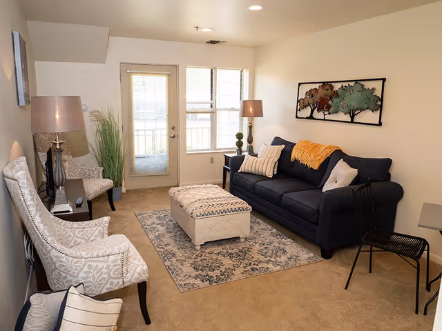 Comfortable living room with a navy couch, two patterned armchairs, an upholstered ottoman on a rug, lamps, wall art, and a door with windows to the outside.