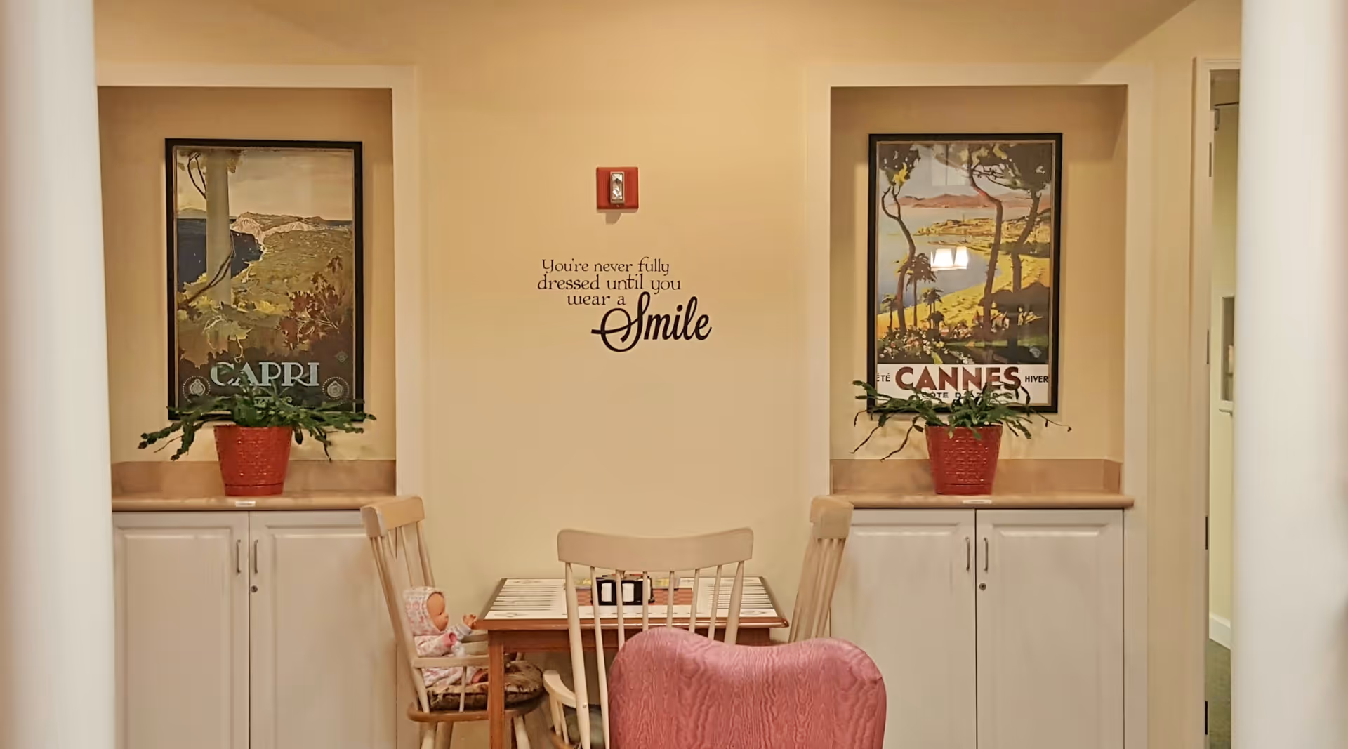 A small dining area with a wooden table and chairs, two framed vintage travel posters on the wall labeled Capri and Cannes, two red potted plants on white cabinets beneath the posters, and a wall decal that reads 'You're never fully dressed until you wear a smile'. A doll is seated on one of the chairs.