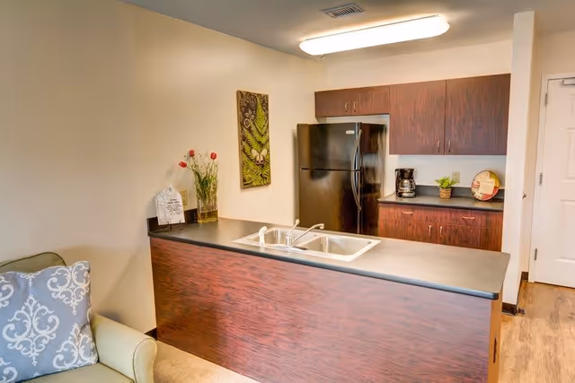 Interior view of a small kitchen area with a dark wood finish. The kitchen features a black refrigerator, upper and lower cabinets, a coffee maker on the counter, and a double sink on a peninsula counter. A piece of wall art and a vase with flowers are visible on the left side. Part of a green armchair with a decorative pillow is seen in the foreground.