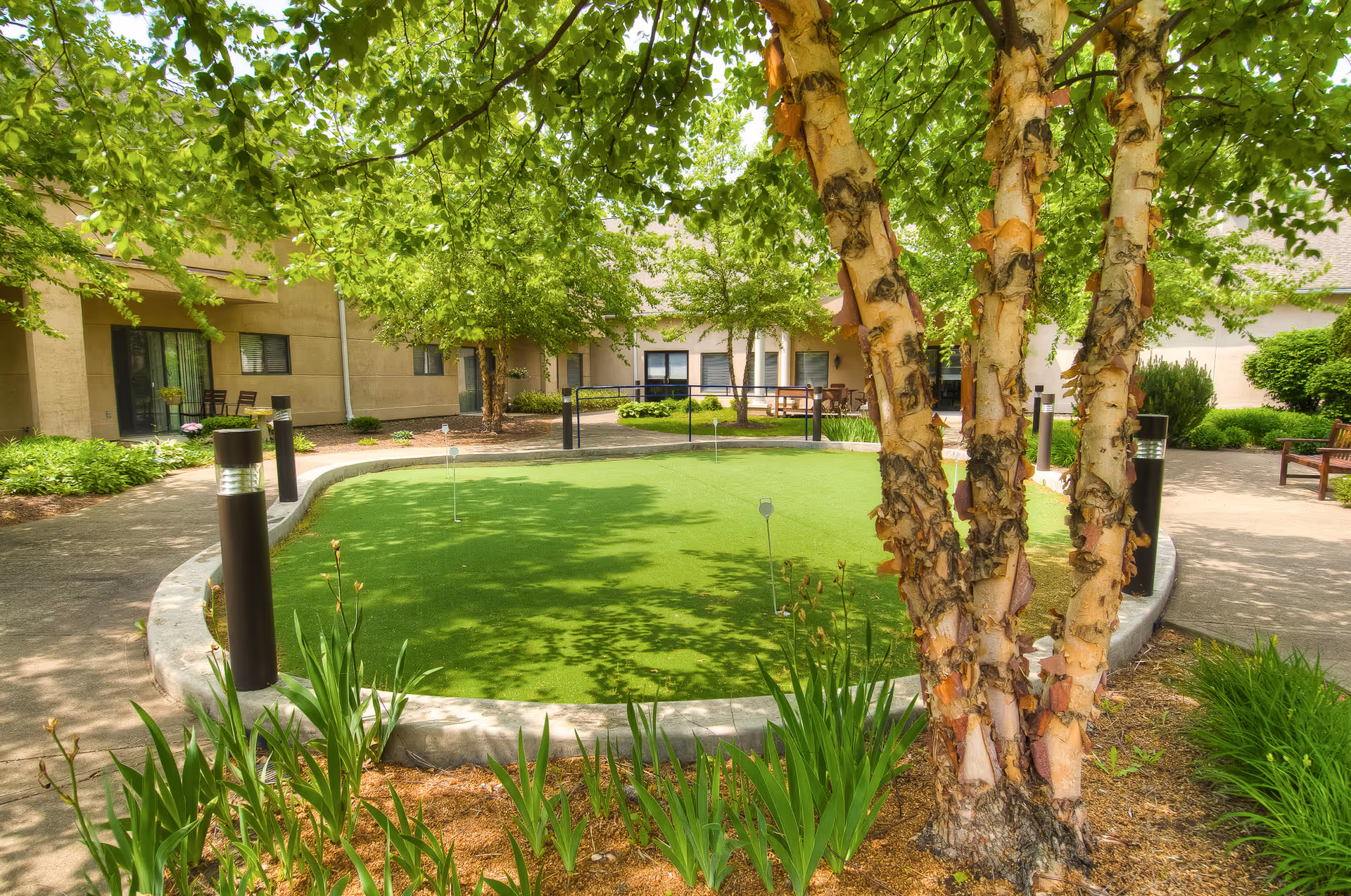 A peaceful outdoor courtyard at The Abbewood featuring a putting green surrounded by a concrete border, lush green trees providing shade, and well-maintained plants and flowers. There are benches and chairs along the building walls, and modern black outdoor lights line the walkway.