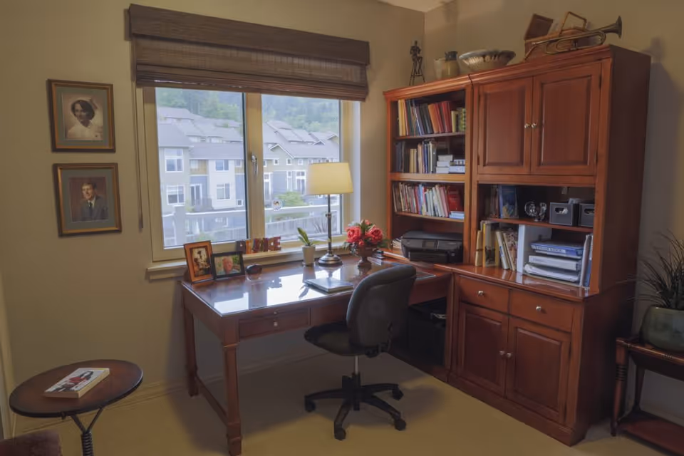 A cozy home office with a wooden desk and matching hutch filled with books and office supplies. A black office chair is positioned at the desk, which holds a lamp, framed photos, a small plant, and a bouquet of flowers. Two framed portraits hang on the wall next to a window with a view of residential buildings outside. A small round side table with a book is also visible.