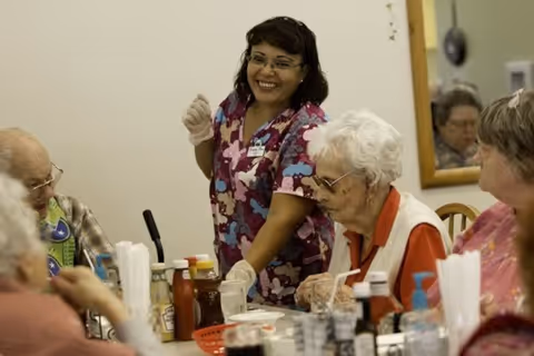 A smiling caregiver wearing a colorful scrub top and gloves interacts with elderly residents seated around a dining table with condiments and utensils in a retirement community dining area.
