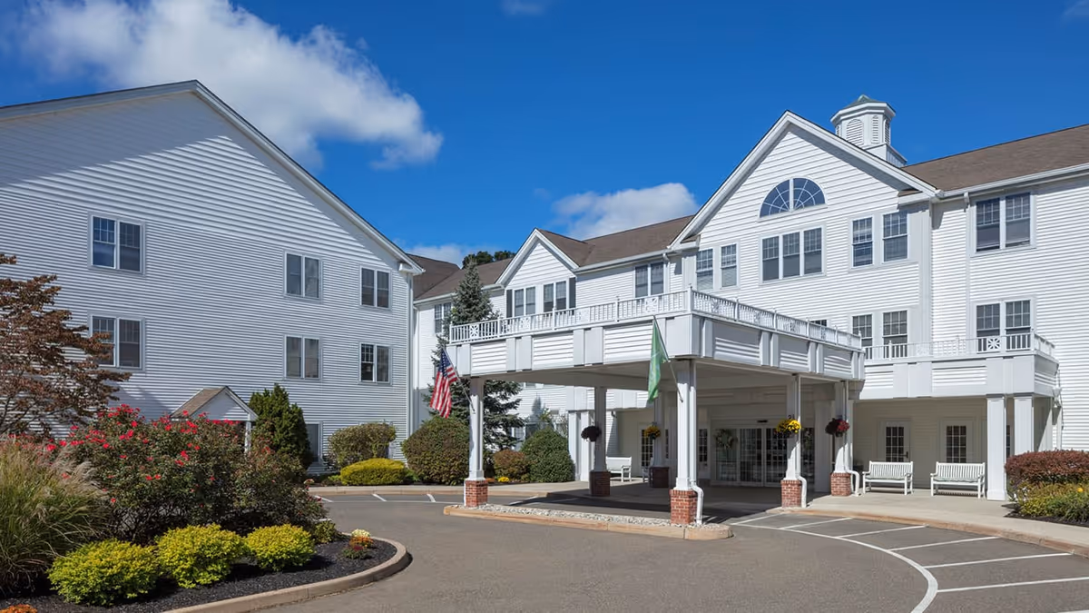 Exterior view of a large white senior living facility building with multiple windows, a covered entrance with white columns and brick bases, surrounded by landscaped bushes and plants under a blue sky with some clouds.