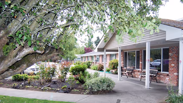 Front exterior of a single-story assisted living building with a covered walkway, benches, and landscaped garden.
