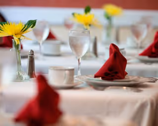 Elegantly set dining tables with water glasses, red folded napkins, white plates, and small vases holding yellow flowers.