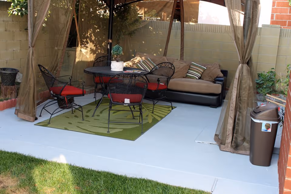 Outdoor patio area with a round black metal table and four matching chairs with red cushions on a green rug. There is a brown cushioned sofa with striped pillows against a brick wall. The area is shaded by a canopy with brown curtains tied to the posts. A brown trash bin is visible on the right side near the brick wall.