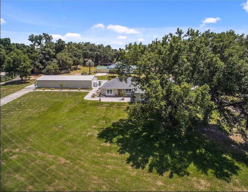 Aerial view of a single-story building with a large green lawn and several trees surrounding it under a blue sky with some clouds.