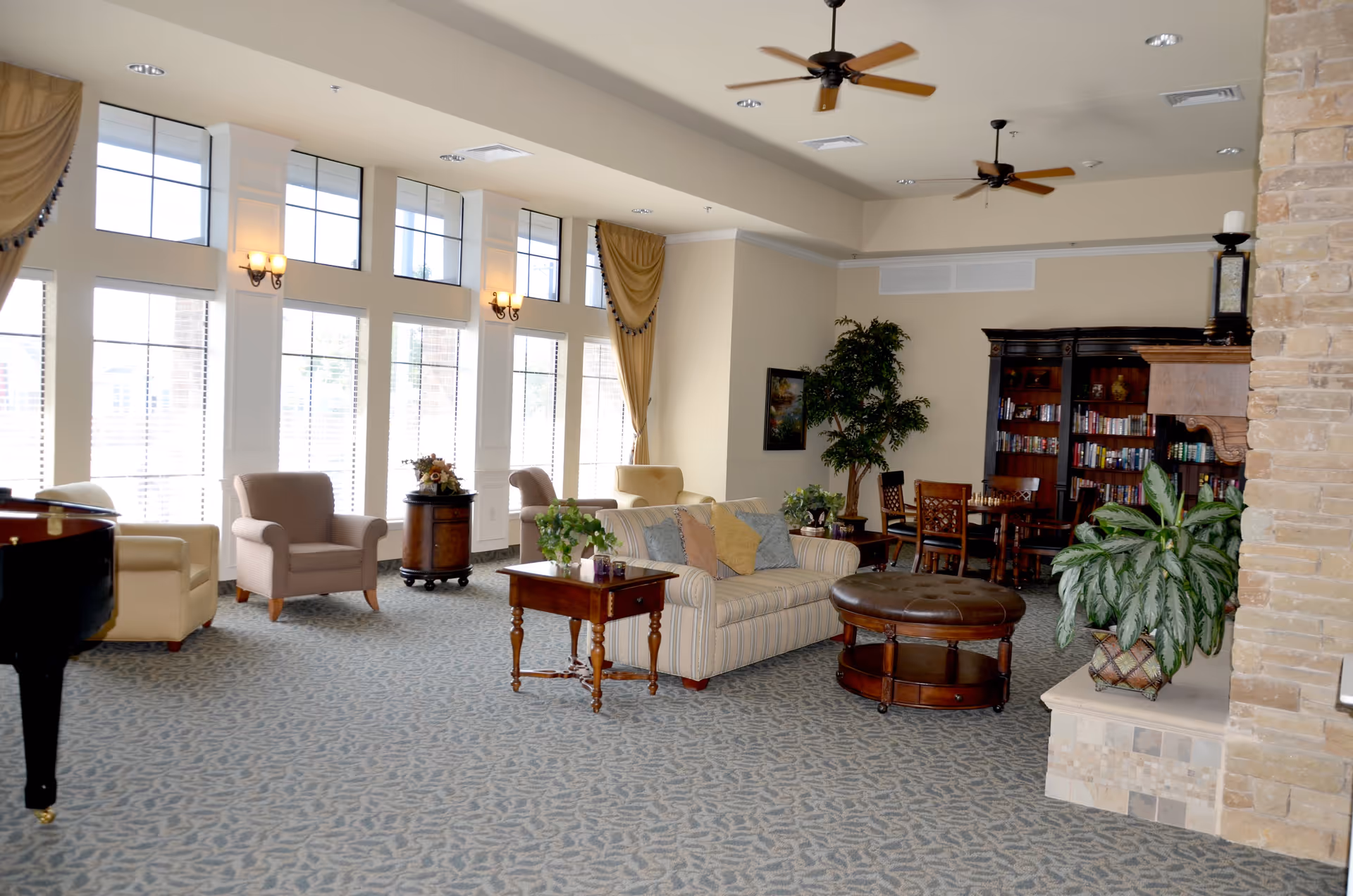 A spacious and well-lit living room area with large windows along one wall, beige walls, and ceiling fans. The room features a striped sofa with colorful pillows, several armchairs, a round leather ottoman, wooden side tables with plants, a large wooden bookshelf filled with books, and a stone fireplace. There are also decorative curtains and wall sconces providing warm lighting.