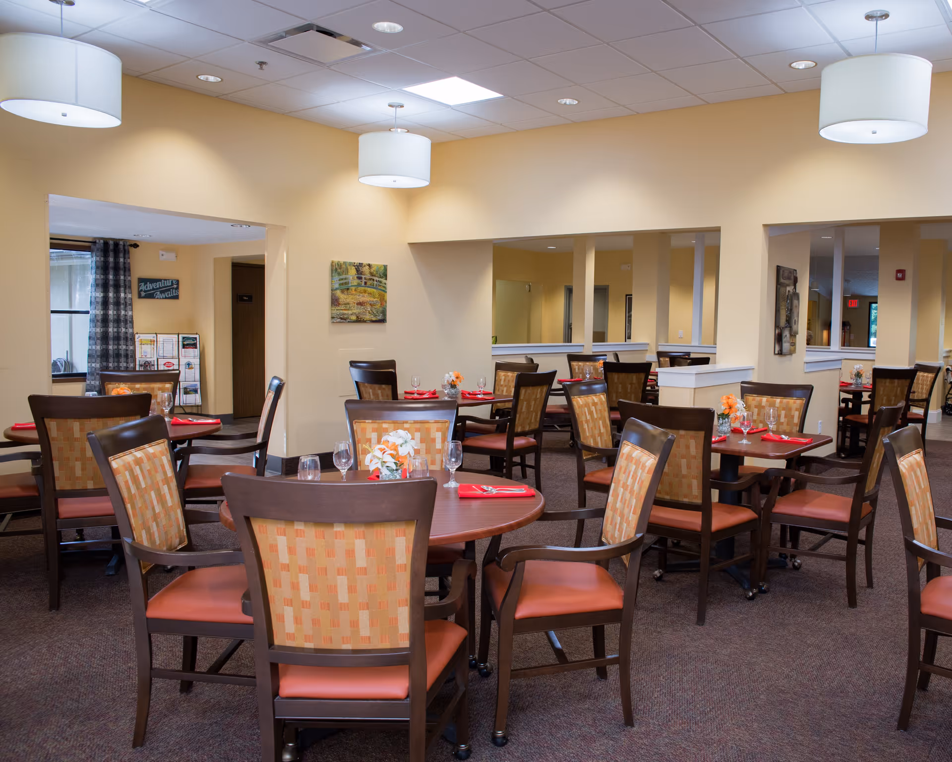 A dining room in Cardinal Retirement Village with multiple round tables set with glasses, napkins, and small flower arrangements. The room has beige walls, carpeted floors, and ceiling lights with white lampshades. There are several wooden chairs with patterned upholstery around the tables. In the background, there are windows with curtains and wall decorations.