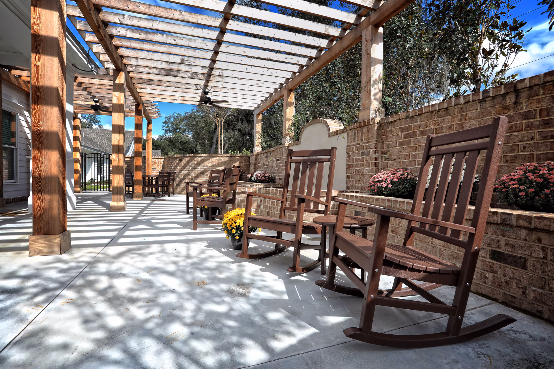 Shaded outdoor patio with wooden rocking chairs under a pergola beside a brick wall and potted flowers.