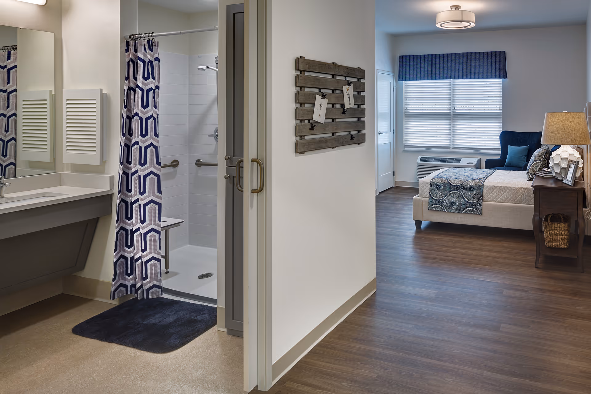 Interior view of a senior living facility room at Walnut Crossing showing a bathroom with a shower area featuring a blue and white patterned curtain and a sink with a large mirror. Adjacent to the bathroom is a bedroom area with a bed, a blue armchair, a window with blinds and a blue valance, a wooden nightstand with a lamp, and a decorative wall piece.
