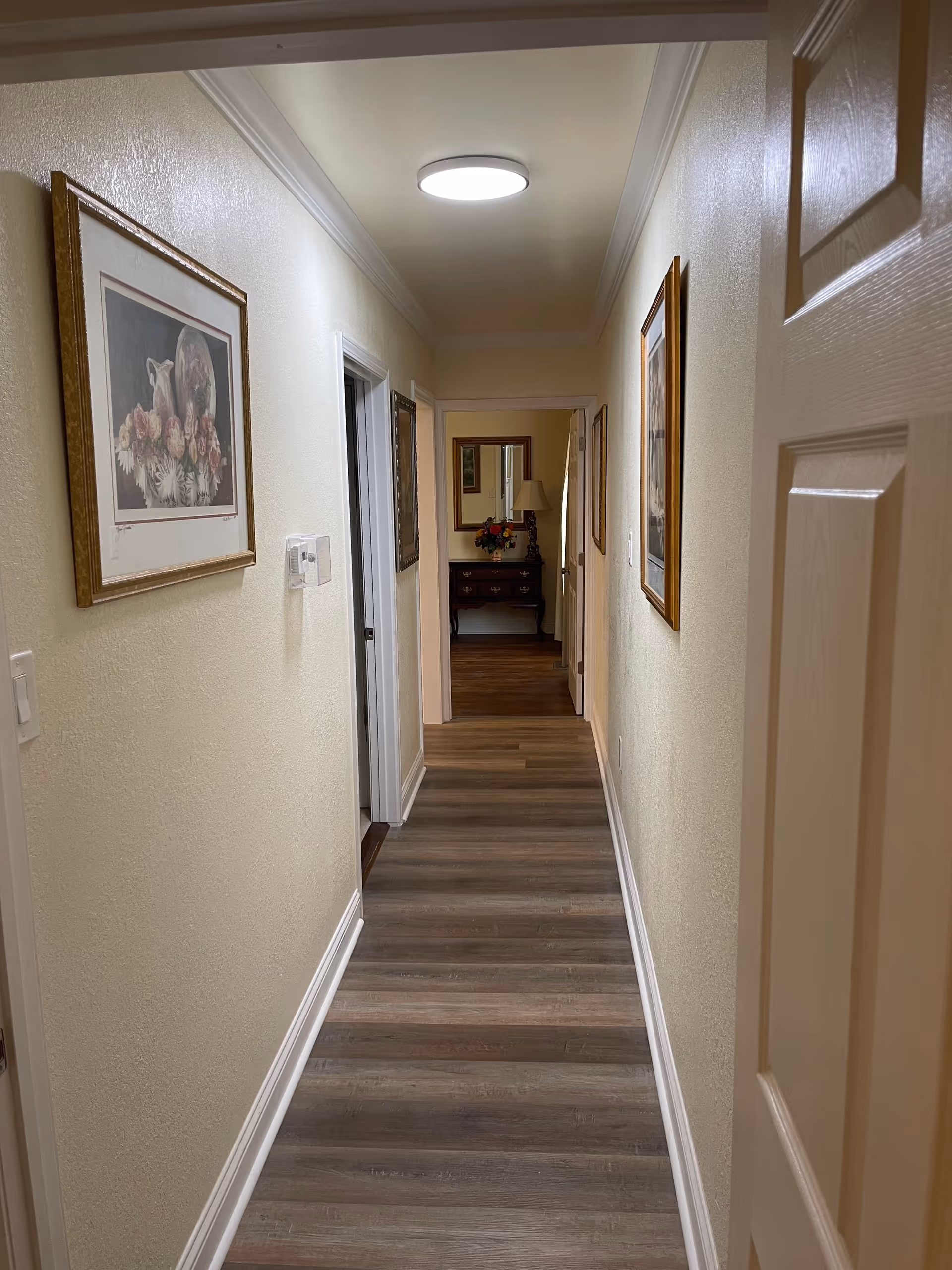 Narrow well-lit hallway with wood-look flooring, framed pictures on the walls, and a table with a lamp and flowers at the far end.