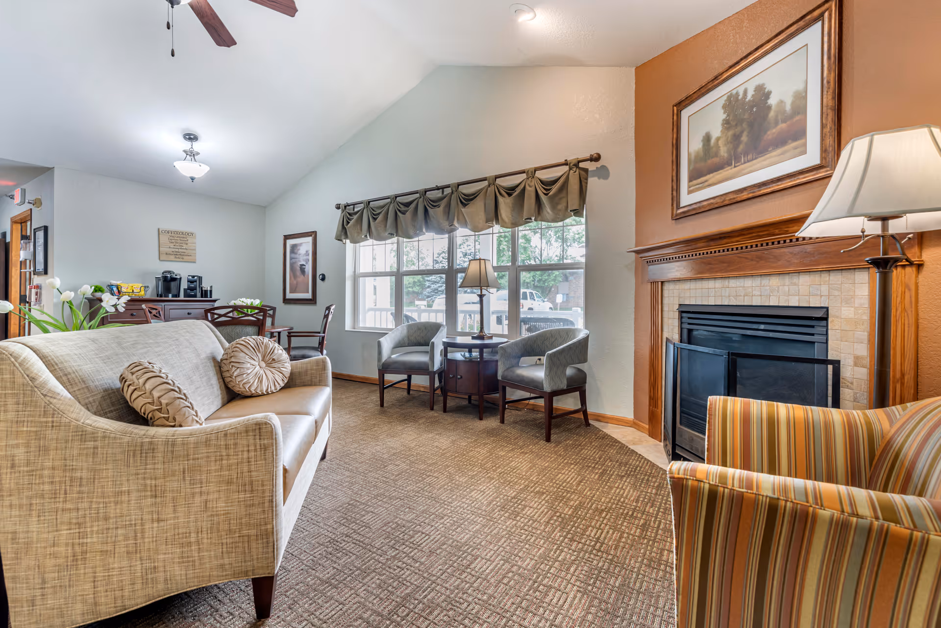 A cozy living room area in a senior living facility with a beige sofa adorned with two round pillows, two gray armchairs around a small wooden table with a lamp, a striped armchair, and a fireplace with a framed landscape painting above it. The room has a large window with a valance, carpeted floor, and a ceiling fan with a light fixture. In the background, there is a small dining table with chairs and a coffee station on a sideboard.