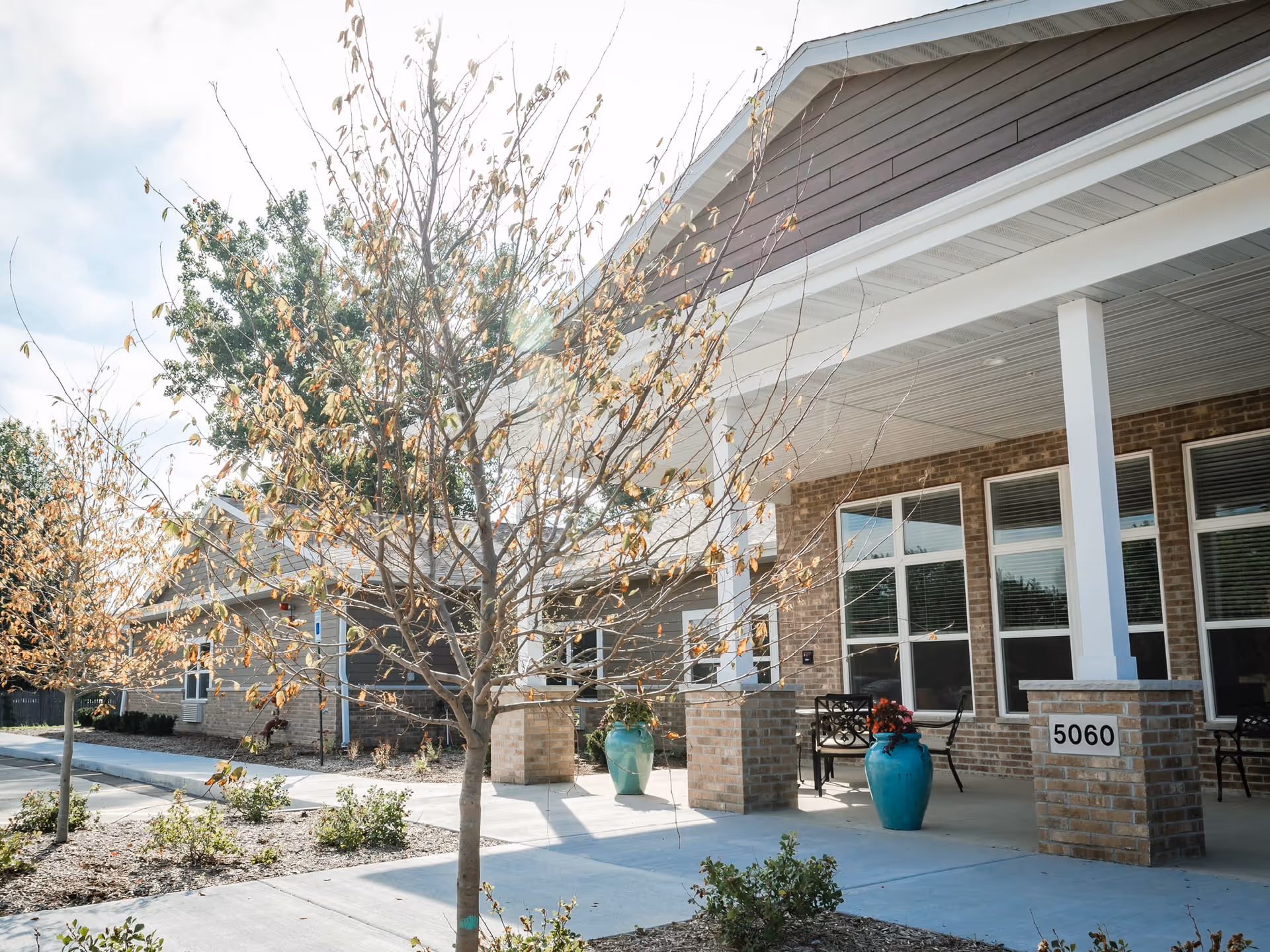 Exterior view of Kenosha Place Senior Living showing a covered entrance with brick pillars, large windows, and a sidewalk lined with small trees and plants. Two large blue planters with flowers are placed near the entrance.