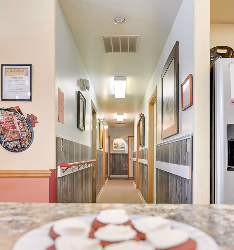 View down a well-lit hallway in an assisted living facility with wood paneling on the lower half of the walls and handrails on both sides. The hallway has several closed doors and framed pictures on the walls. In the foreground, there is a blurred plate of cookies on a countertop, and part of a refrigerator is visible on the right side.