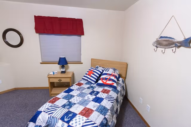 A small bedroom with a single bed covered in a nautical-themed quilt and pillows featuring anchors and stars. Next to the bed is a wooden nightstand with a blue lamp and a small item on it. The window behind the nightstand has a red valance and closed blinds. A round mirror hangs on the left wall, and a decorative fish with hooks is mounted on the right wall. The room has light-colored walls and carpeted flooring.