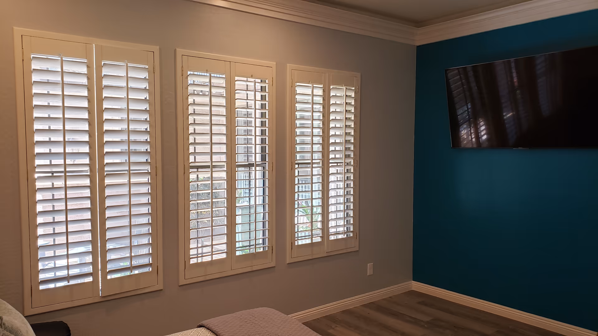 A room with four large windows covered by white plantation shutters. The walls are painted light gray with one accent wall painted teal. A flat-screen TV is mounted on the teal wall. The floor is wood with a light finish.