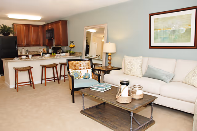 A cozy living room and kitchen area in a senior living facility. The living room features a beige sofa with decorative pillows, a wooden coffee table with candles and books, a chair with a floral cushion, a side table with a lamp, and a large mirror leaning against the wall. The kitchen has wooden cabinets, a black refrigerator, a microwave, and a counter with three wooden stools.