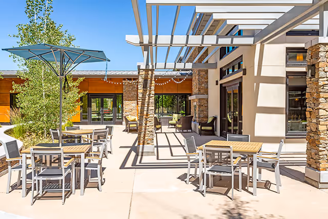 Outdoor patio area at Sagewood at Daybreak with several tables and chairs arranged for seating. There is a large umbrella providing shade over one table, and a pergola casting shadows on the ground. The building features stone and wood accents with large windows and doors, and there are trees and greenery around the patio.