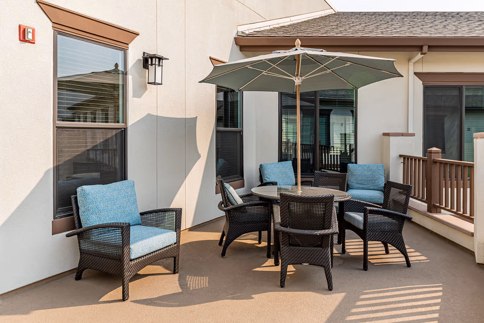 Outdoor patio area with a round glass table surrounded by six wicker chairs with blue cushions and a large green umbrella providing shade. The patio is adjacent to a building with beige walls and several windows.