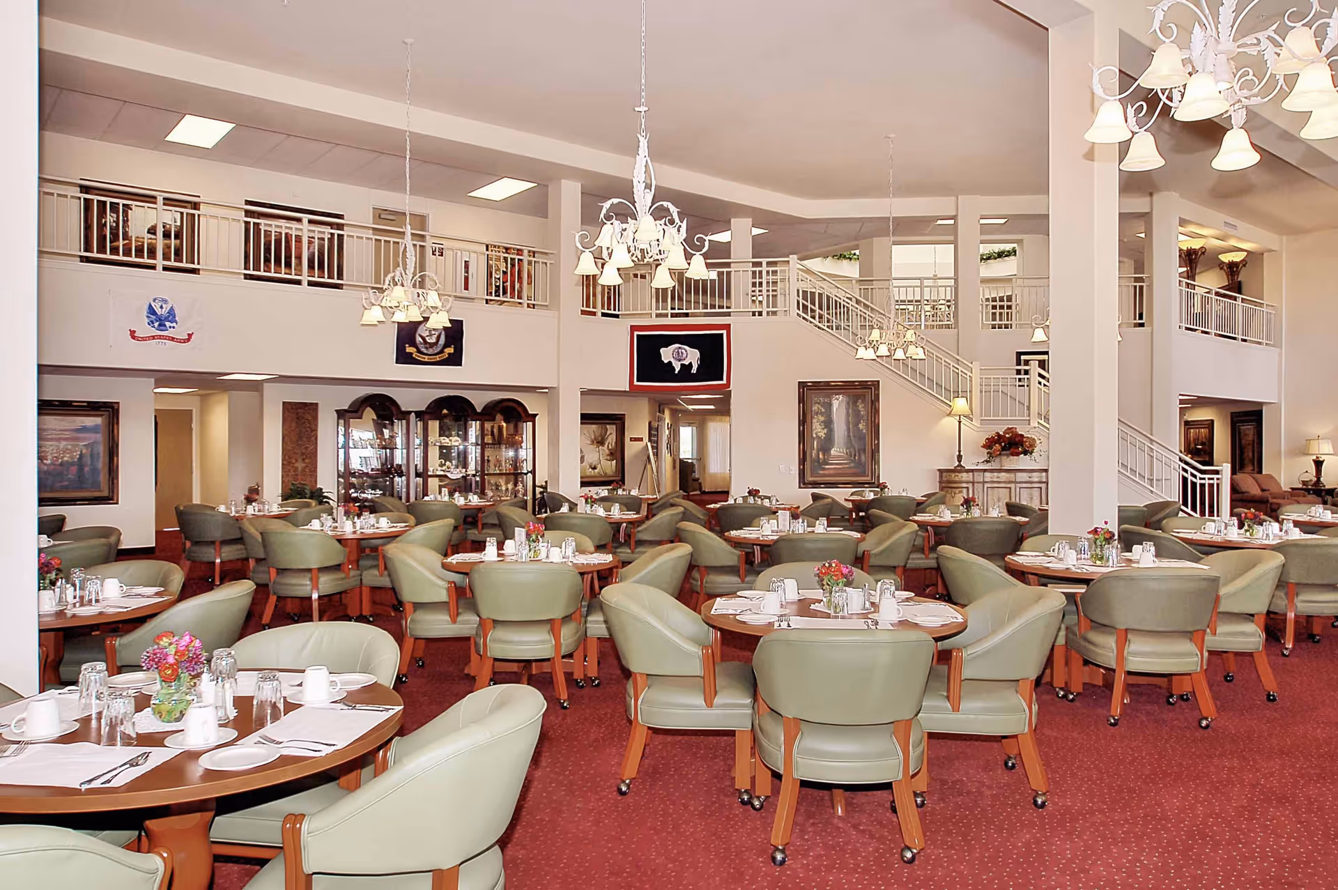 A spacious dining room with multiple round tables set with white cups, glasses, plates, and silverware. Each table has green cushioned chairs with wooden legs on a red carpeted floor. The room features high ceilings with white chandeliers, a staircase leading to an upper level with railings, and various framed artworks and flags on the walls.