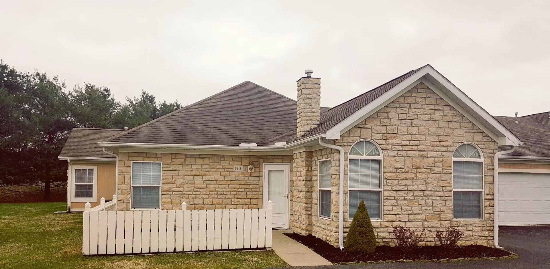 Front exterior of a single-story stone building with a white picket fence, chimney, and attached garage.