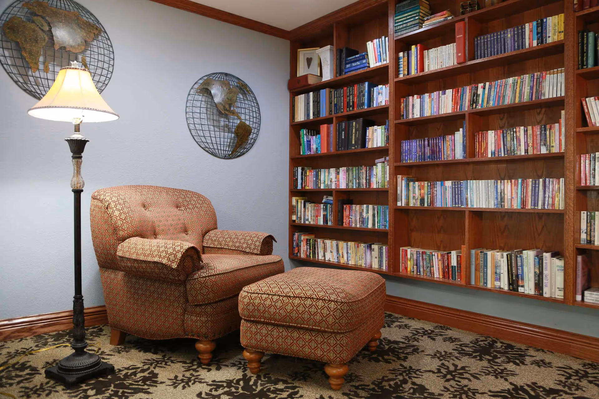 Cozy reading nook with a patterned armchair and matching ottoman next to a floor lamp, set against a wall with two metal world map decorations and a large wooden bookshelf filled with books.