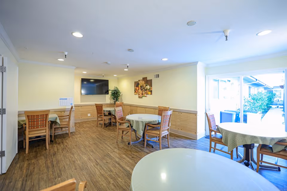 A bright dining room in a senior living facility with round and square tables covered with green tablecloths, wooden chairs, a wall-mounted flat screen TV, a plant, and a painting on the wall. Large windows on the right side let in natural light and show an outdoor walkway with greenery.