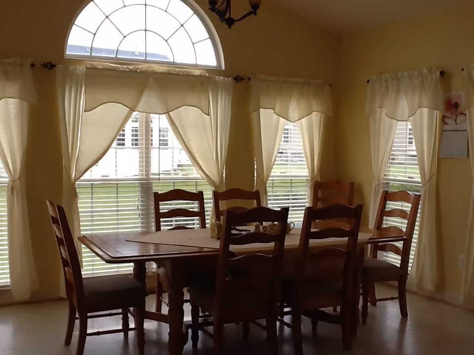 A dining room with a wooden table and six matching wooden chairs. The room has large windows with white curtains and blinds, allowing natural light to enter. The walls are painted a soft yellow color.