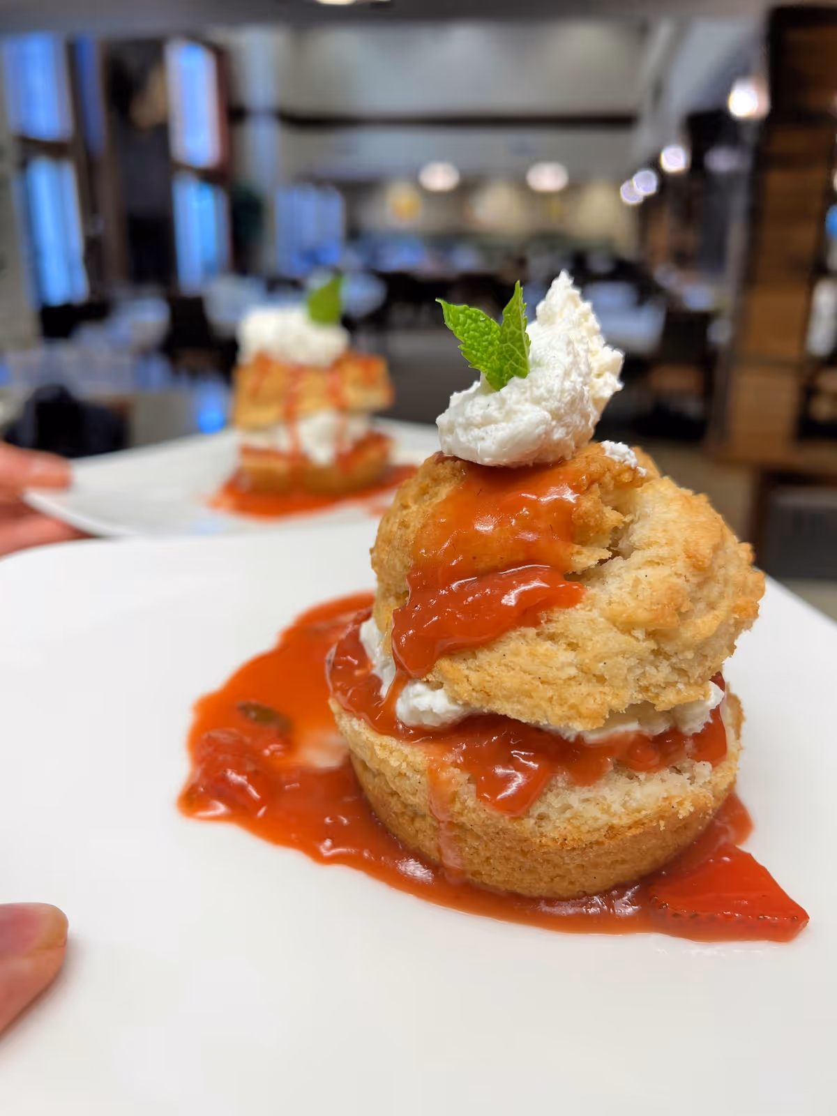 Close-up of a dessert on a white plate featuring a biscuit topped with whipped cream, strawberry sauce, and a small mint leaf, with a blurred background showing a dining area.