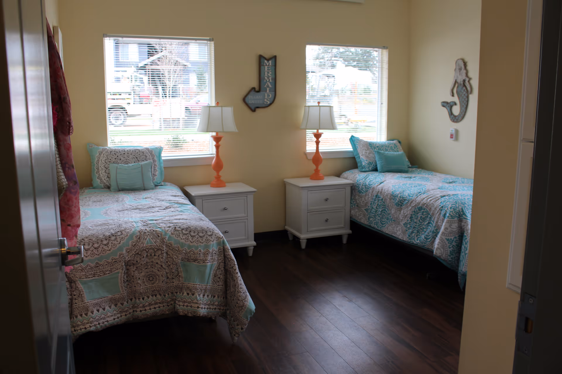 A bedroom with two twin beds, each with patterned blue and beige bedding. Between the beds are two white nightstands with orange lamps. The room has two windows with blinds, and there are decorative wall hangings including a mermaid figure and a sign that says 'MERMAIDS'. The floor is dark wood.